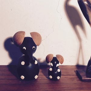 Puede incluir: Tres figuritas de animales decorativas: una garza alta, marrón y blanca, un elefante grande con manchas blancas y negras, y un elefante más pequeño con manchas blancas y negras. Las figuritas están sobre una superficie de madera.