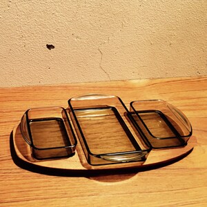 May include: A wooden serving tray with three clear glass dishes. The tray is oval shaped and the dishes are rectangular. The tray is on a wooden surface.