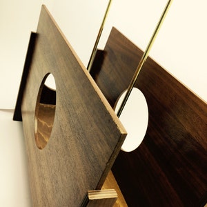 May include: A wooden record holder with a geometric design. The holder is made of dark brown wood with a gold-colored metal handle. The holder has a circular cutout on the front and is designed to hold vinyl records.