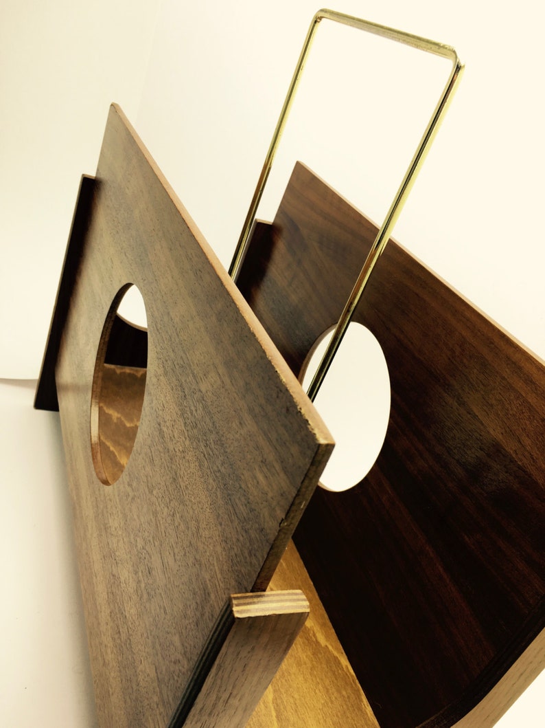 May include: A wooden record holder with a geometric design. The holder is made of dark brown wood with a gold-colored metal handle. The holder has a circular cutout on the front and is designed to hold vinyl records.
