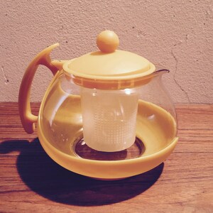 Puede incluir: Una tetera de vidrio amarilla con un colador incorporado. La tetera tiene un cuerpo redondo y un asa curva.