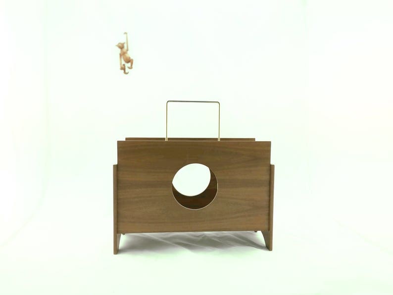 May include: A mid-century modern wooden box with a circular opening and a gold-colored handle. A small monkey figurine hangs from a thin rod above the box. The box is a warm brown color.