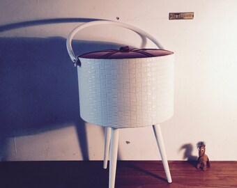May include: White wicker storage basket with a brown lid and four white legs. The basket is sitting on a wooden floor.