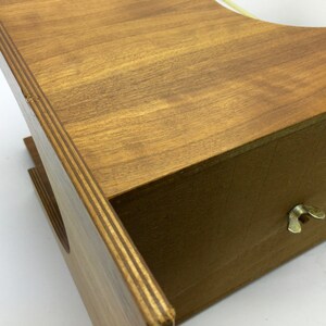 May include: A wooden box with a light brown finish. The box has a drawer with a brass wing nut and a circular opening on top. The wood grain is visible, and the box appears to be handmade.