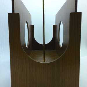 May include: A wooden magazine rack with a mid-century modern design. The rack is made of light brown wood and features two vertical panels with circular cutouts. A brass rod runs through the center. The rack is approximately 20 cm wide.