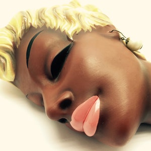 Puede incluir: Una escultura de pared de cerámica de la cara de una mujer con los ojos cerrados y la boca abierta de color rosa. La mujer tiene el pelo rubio y lleva un solo pendiente de perla.