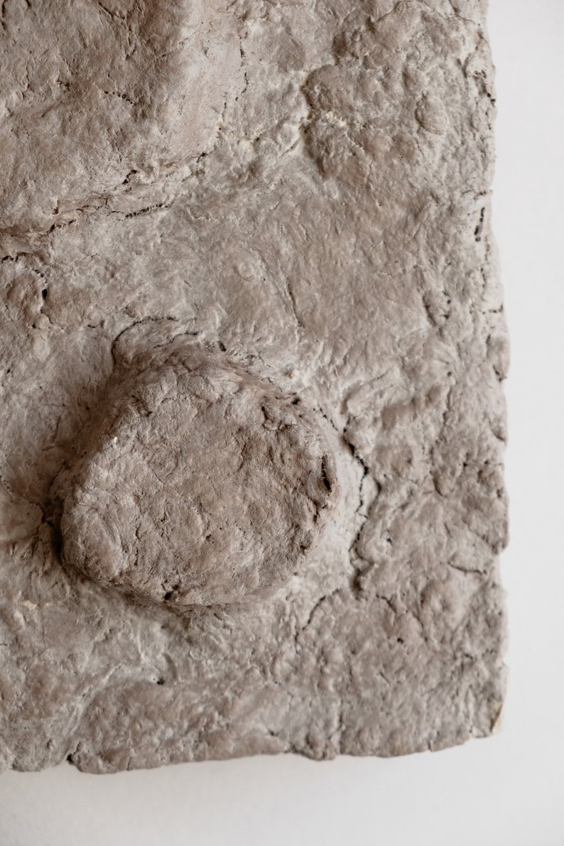 Puede incluir: Primer plano de una superficie rectangular texturizada, gris-marr&oacute;n, con un elemento redondeado en relieve. La superficie tiene un aspecto rugoso e irregular con grietas y variaciones en la textura, lo que sugiere un material natural o hecho a mano.
