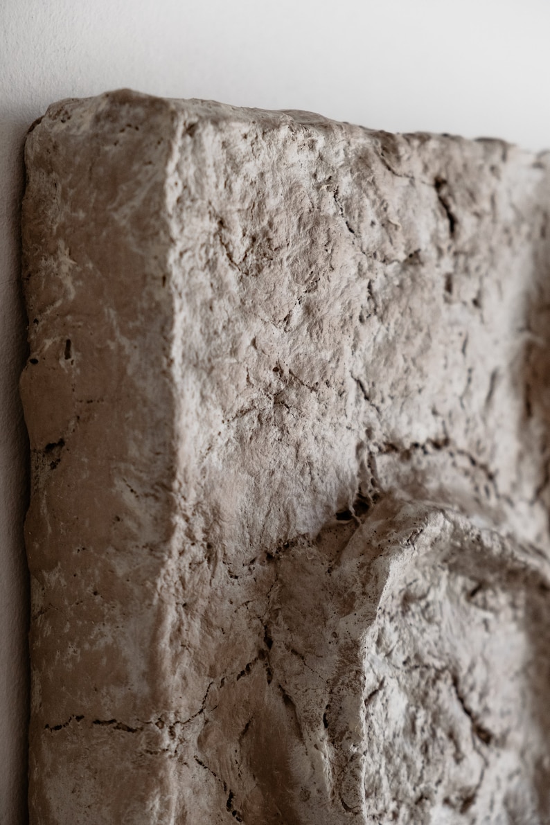 Puede incluir: Primer plano de una obra de arte mural rectangular marr&oacute;n claro texturizada. La superficie tiene una textura rugosa e irregular con grietas visibles y variaciones de color, creando una est&eacute;tica natural y terrosa. La obra de arte est&aacute; montada en una pared blanca.