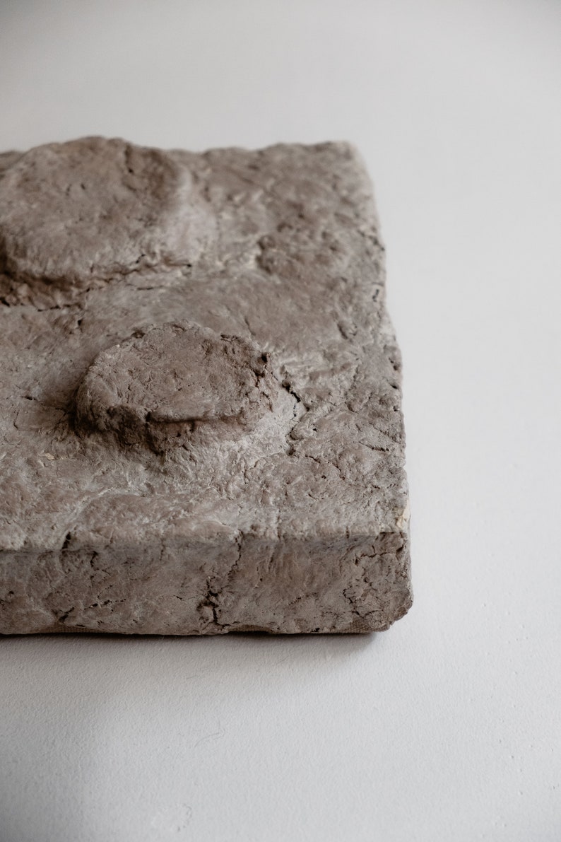 Puede incluir: Un bloque rectangular gris texturizado con una superficie rugosa y protuberancias redondeadas. El bloque tiene un aspecto natural y terroso, con grietas e imperfecciones visibles. El objeto es probablemente una pieza decorativa o un material de construcci&oacute;n.