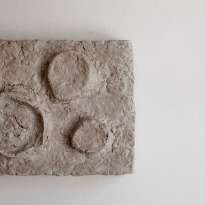 Puede incluir: Una pieza de arte mural rectangular y texturizada, de color marr&oacute;n claro, con tres formas circulares en relieve. La obra de arte tiene una superficie rugosa y desigual, sobre un fondo blanco.
