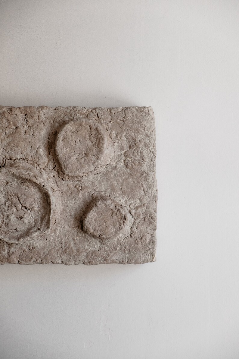 Puede incluir: Una pieza de arte mural rectangular y texturizada, de color marr&oacute;n claro, con tres formas circulares en relieve. La obra de arte tiene una superficie rugosa y desigual, sobre un fondo blanco.