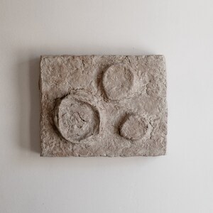 Puede incluir: Una pieza de arte mural rectangular y texturizada de color beige con tres hendiduras circulares. El c&iacute;rculo m&aacute;s grande tiene un interior agrietado y texturizado. La obra de arte est&aacute; montada en una pared blanca.