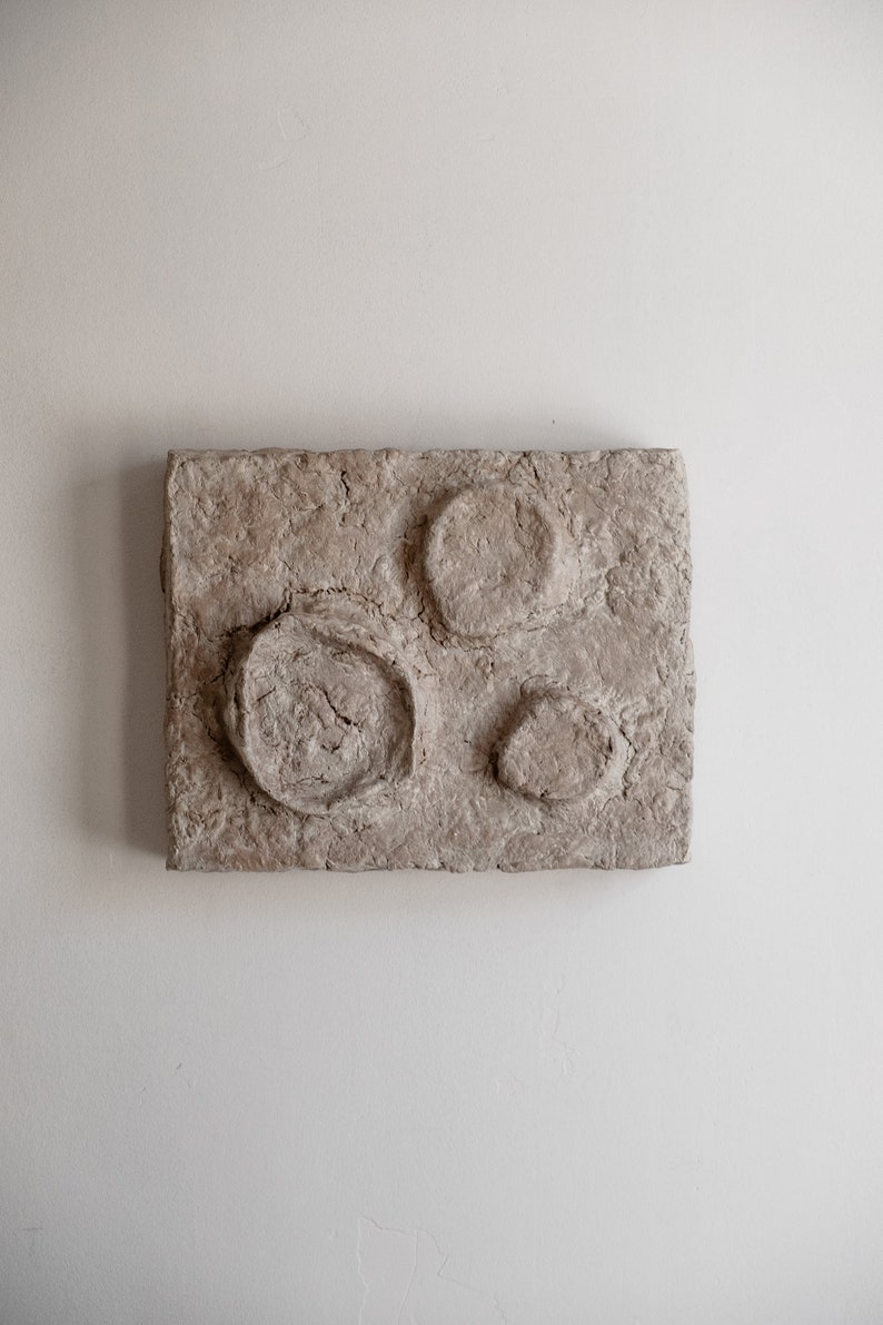 Puede incluir: Una pieza de arte mural rectangular y texturizada de color beige con tres hendiduras circulares. El c&iacute;rculo m&aacute;s grande tiene un interior agrietado y texturizado. La obra de arte est&aacute; montada en una pared blanca.