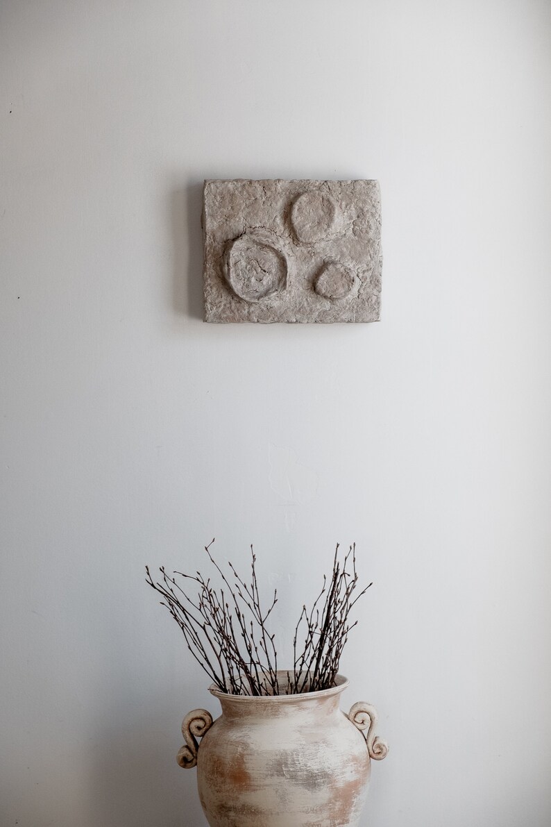 Puede incluir: Una obra de arte mural texturizada de tonos neutros con tres dise&ntilde;os circulares en relieve cuelga sobre un jarr&oacute;n decorativo blanquecino. El jarr&oacute;n est&aacute; lleno de ramas delgadas de color marr&oacute;n oscuro, creando una est&eacute;tica minimalista.