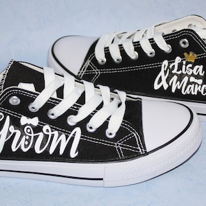 Könnte beinhalten: Ein Paar schwarze Leinenschuhe mit weißen Schnürsenkeln und Sohlen. Auf einem Schuh steht "Groom" in weißer Schrift mit einer Fliege, auf dem anderen "Lisa & Marcel" mit einer goldenen Krone. Schuhe zum Thema Hochzeit.