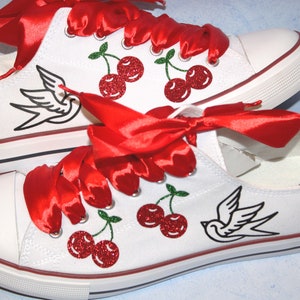 Könnte beinhalten: Weiße Canvas-Sneaker mit niedrigem Schaft und roten Satin-Schnürsenkeln. Die Schuhe sind mit gestickten Kirschen und einem Schwalben-Tattoo-Design verziert.
