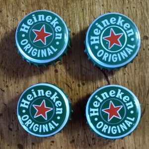 May include: Four green and silver Heineken bottle caps. Each cap features the Heineken logo with a red star in the center, surrounded by the words "Heineken" and "Original" in white. The caps are arranged in a square.