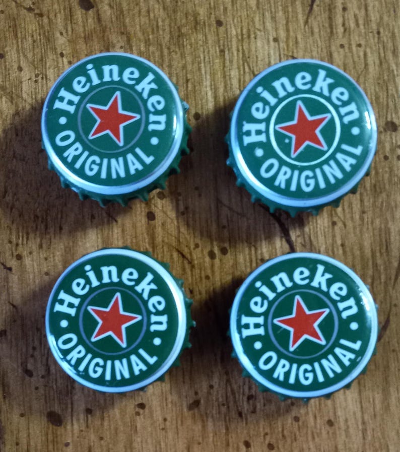 May include: Four green and silver Heineken bottle caps. Each cap features the Heineken logo with a red star in the center, surrounded by the words "Heineken" and "Original" in white. The caps are arranged in a square.