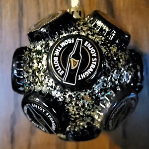 Puede incluir: Adorno navide&ntilde;o negro y dorado hecho de tapas de botellas con el texto "Enjoy Straight From The Bottle" y una silueta de una botella de cerveza.