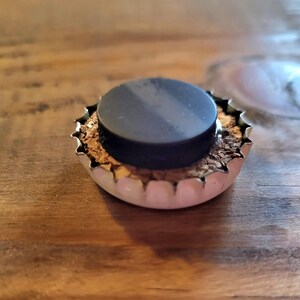 May include: A bottle cap magnet with a black circular magnet adhered to the center. The cap is silver with a jagged edge and a cork-like material visible inside. The magnet is on a wooden surface.