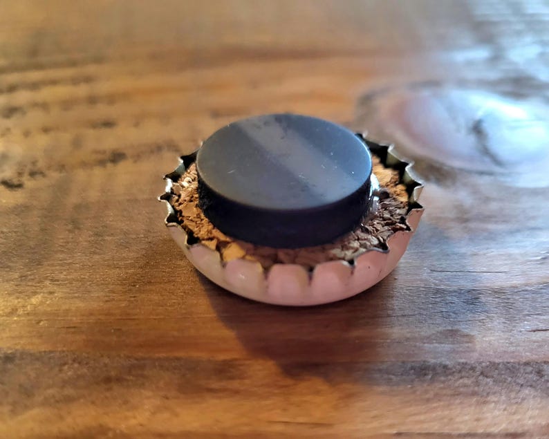 May include: A bottle cap magnet with a black circular magnet adhered to the center. The cap is silver with a jagged edge and a cork-like material visible inside. The magnet is on a wooden surface.