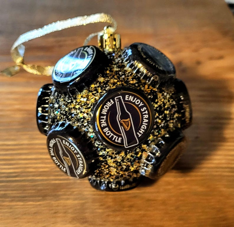 Puede incluir: Un adorno navide&ntilde;o negro y dorado hecho de tapas de botellas. El adorno presenta un gr&aacute;fico en blanco y negro de una botella de cerveza con el texto "Enjoy Straight From The Bottle".