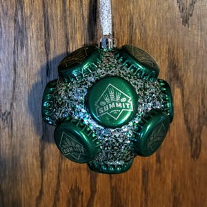 May include: Green Christmas ornament made from bottle caps with silver glitter. The ornament features the Summit Brewing Co. logo.
