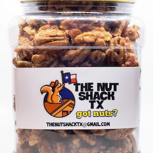 Puede incluir: Un tarro de pl&aacute;stico transparente lleno de nueces pecanas confitadas. El tarro tiene una tapa amarilla y una etiqueta blanca con el texto "THE NUT SHACK TX got nuts?" y un logotipo de una ardilla. Tambi&eacute;n es visible la direcci&oacute;n de correo electr&oacute;nico thenutshacktx@gmail.com.