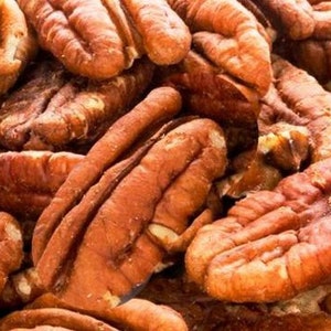 K&ouml;nnte beinhalten: Nahaufnahme eines Haufens Pekannussh&auml;lften. Die N&uuml;sse haben eine warme braune Farbe mit strukturierten Oberfl&auml;chen und sichtbaren Rippen. Das Bild hebt die nat&uuml;rlichen Formen und Details der Pekann&uuml;sse hervor und zeigt ihre essbare Natur.