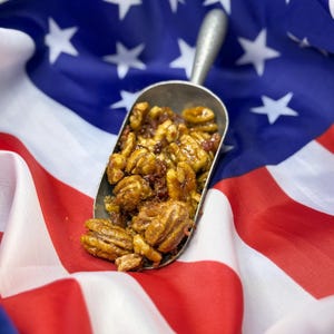 Puede incluir: Una cuchara met&aacute;lica llena de nueces pecanas glaseadas y otras nueces, sobre una bandera estadounidense. Las nueces son una mezcla de colores marr&oacute;n y dorado, con un glaseado brillante. La bandera roja y blanca y el campo azul estrellado proporcionan un fondo patri&oacute;tico.