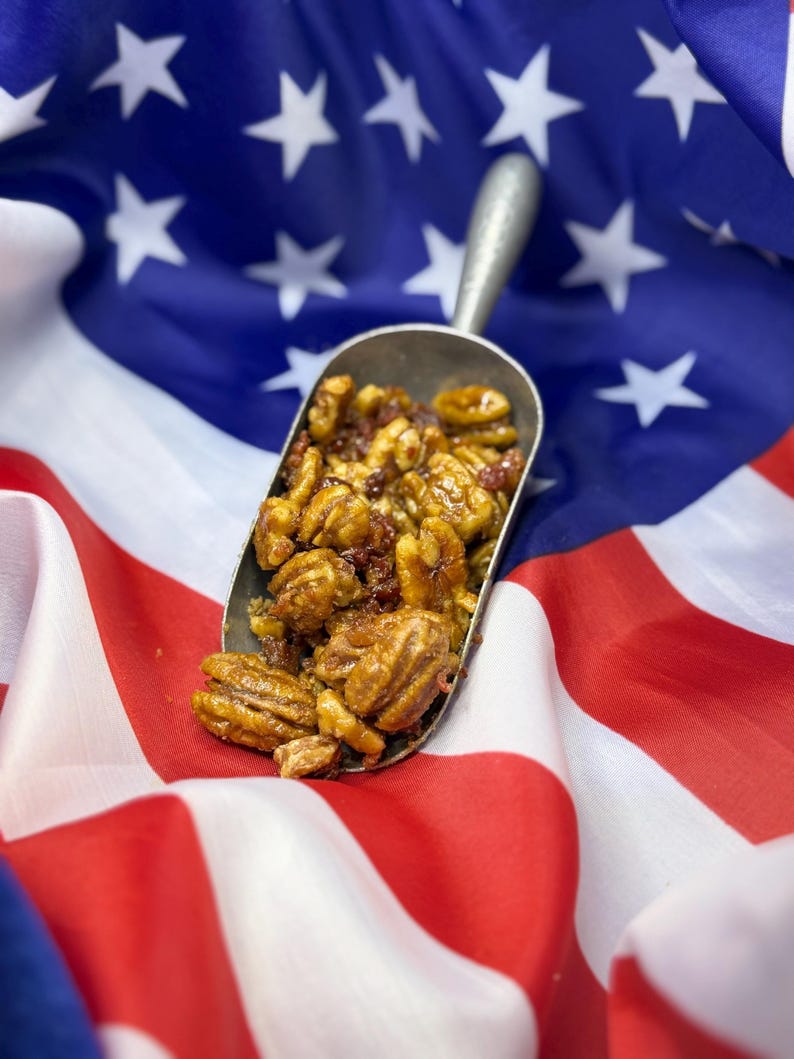Puede incluir: Una cuchara met&aacute;lica llena de nueces pecanas glaseadas y otras nueces, sobre una bandera estadounidense. Las nueces son una mezcla de colores marr&oacute;n y dorado, con un glaseado brillante. La bandera roja y blanca y el campo azul estrellado proporcionan un fondo patri&oacute;tico.