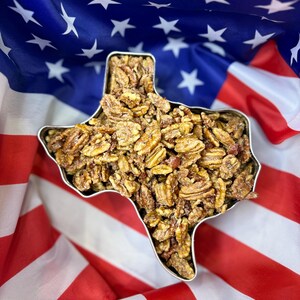 Puede incluir: Un recipiente met&aacute;lico con la forma del estado de Texas lleno de nueces pecanas confitadas. El recipiente se encuentra sobre un fondo de la bandera estadounidense, con rayas rojas y blancas y un campo azul de estrellas blancas.