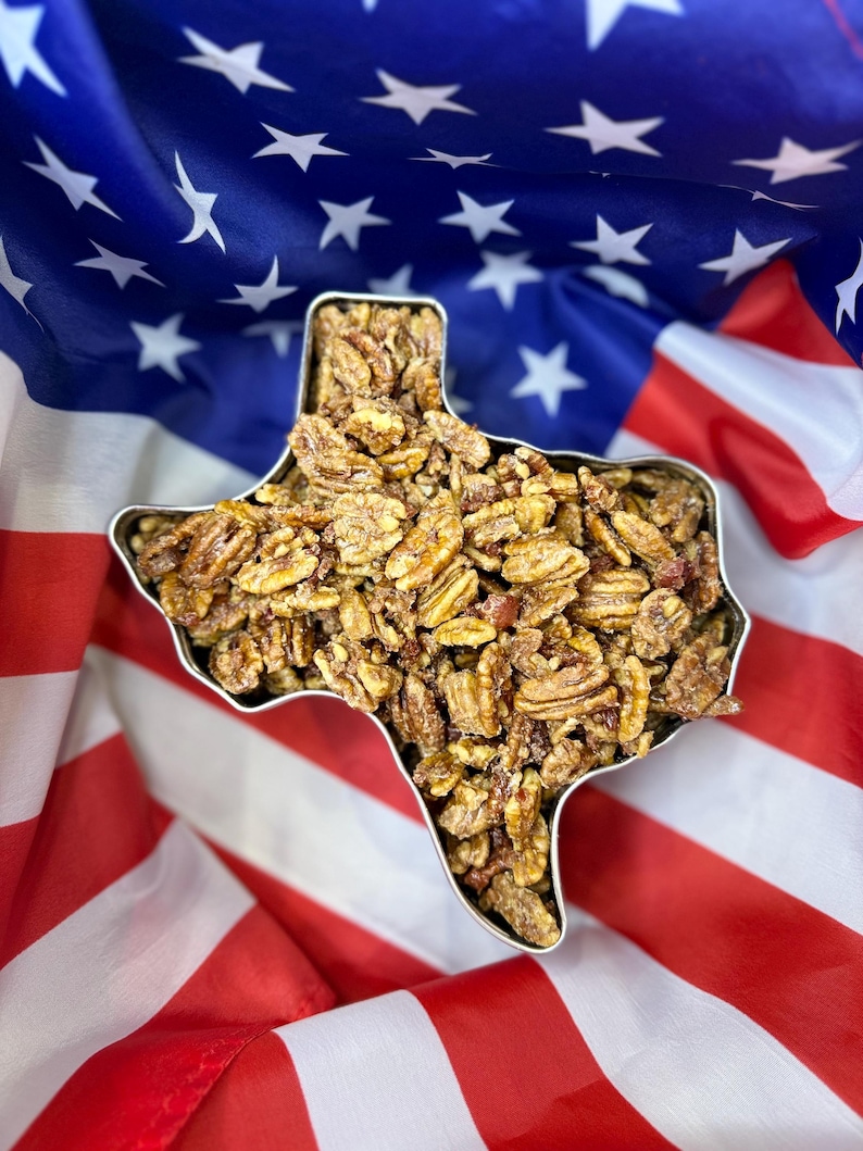 Puede incluir: Un recipiente met&aacute;lico con la forma del estado de Texas lleno de nueces pecanas confitadas. El recipiente se encuentra sobre un fondo de la bandera estadounidense, con rayas rojas y blancas y un campo azul de estrellas blancas.