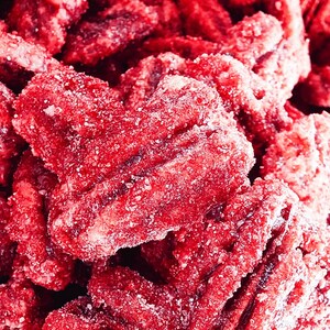 K&ouml;nnte beinhalten: Nahaufnahme von gezuckerten Pekann&uuml;ssen. Das Bild zeigt einen Haufen hellroter, herzf&ouml;rmiger Pekann&uuml;sse, die mit wei&szlig;en Zuckerkristallen bedeckt sind. Die N&uuml;sse haben eine strukturierte Oberfl&auml;che und sind eng aneinandergereiht.