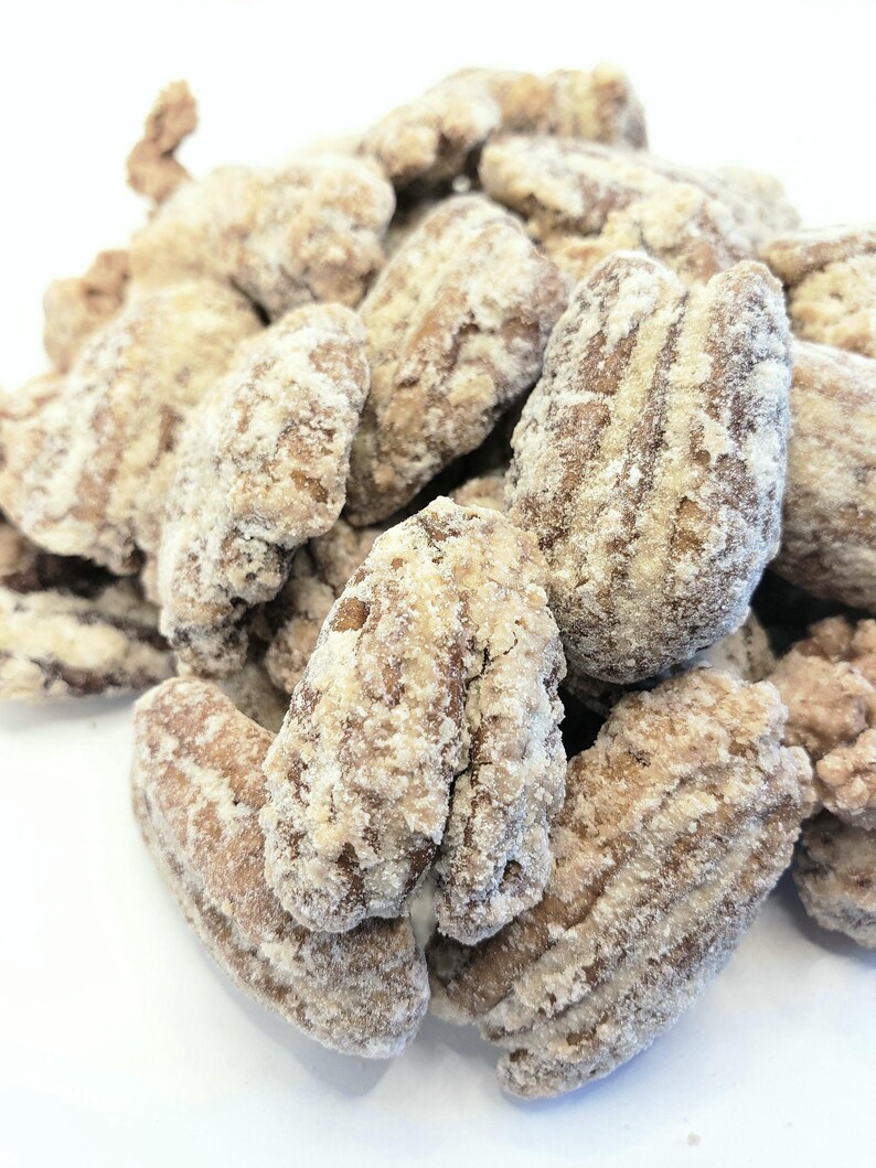 K&ouml;nnte beinhalten: Nahaufnahme eines Haufens gezuckerter Pekann&uuml;sse. Die Pekann&uuml;sse sind hellbraun mit einer wei&szlig;en, zuckerhaltigen Beschichtung. Die N&uuml;sse sind zusammengeballt, wobei einige einzelne N&uuml;sse sichtbar sind. Der Hintergrund ist schlicht wei&szlig;.