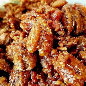 May include: Close-up of candied pecans, showcasing a glossy, caramelized coating. The nuts are a rich brown color, with a slightly textured surface. The image highlights the details of the nuts, suggesting a sweet treat or snack.