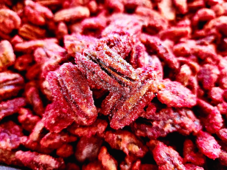Puede incluir: Primer plano de nueces pecanas confitadas, mostrando un color marr&oacute;n rojizo intenso con un recubrimiento azucarado. Las nueces est&aacute;n dispuestas juntas, llenando el encuadre. La imagen resalta la textura y el dulzor del snack.