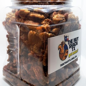Puede incluir: Un tarro de pl&aacute;stico transparente lleno de nueces pecanas confitadas. El tarro tiene forma rectangular y una tapa transparente. Una etiqueta blanca en el tarro dice "THE NUT SHACK TX got nuts?" con un logotipo de ardilla.