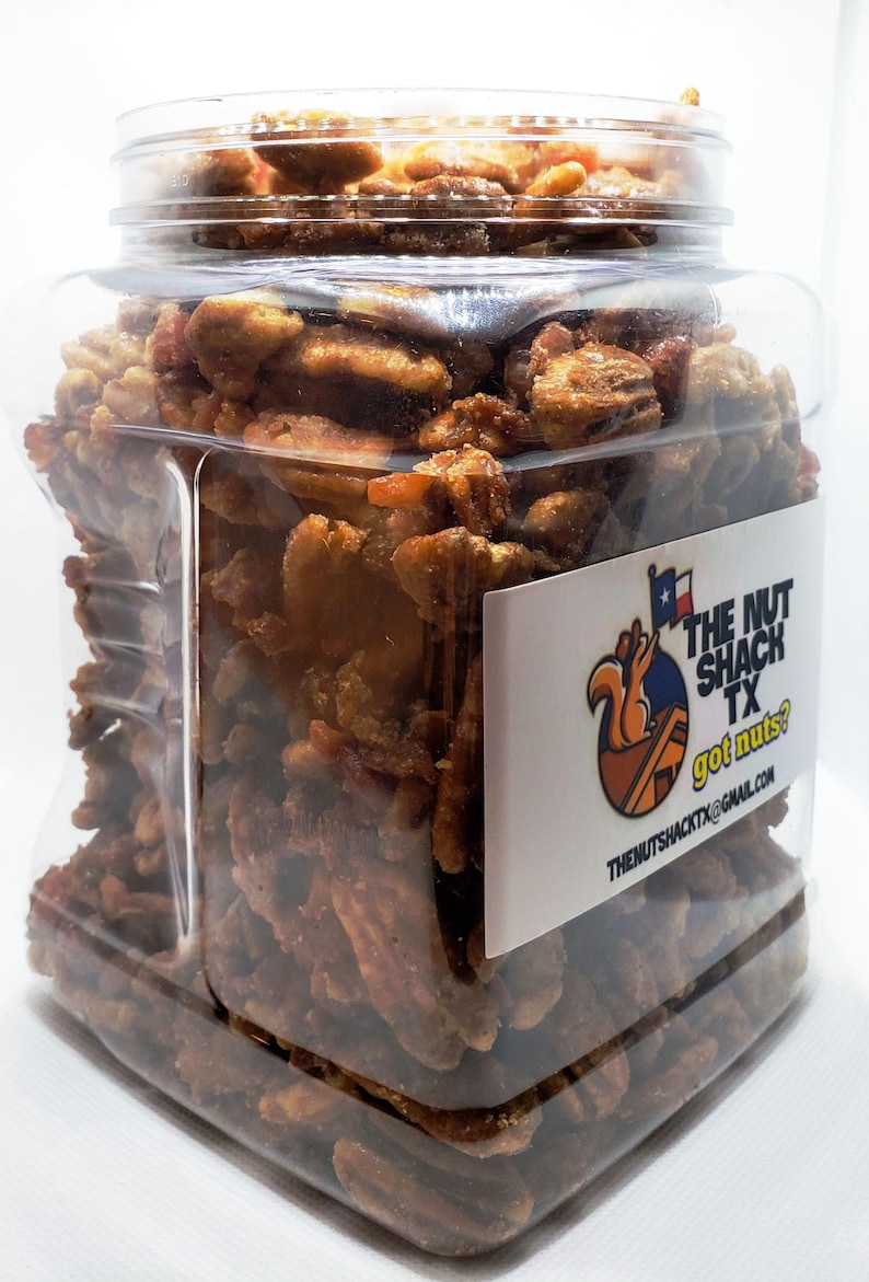 Puede incluir: Un tarro de pl&aacute;stico transparente lleno de nueces pecanas confitadas. El tarro tiene forma rectangular y una tapa transparente. Una etiqueta blanca en el tarro dice "THE NUT SHACK TX got nuts?" con un logotipo de ardilla.