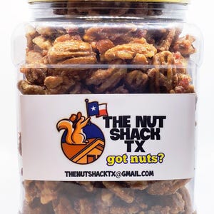K&ouml;nnte beinhalten: Ein durchsichtiges Plastikglas gef&uuml;llt mit Pekannussh&auml;lften, bedeckt mit einem gelben Deckel. Das Glas hat ein wei&szlig;es Etikett mit dem Text "THE NUT SHACK TX got nuts?" und einem Logo eines Eichh&ouml;rnchens, das eine Flagge h&auml;lt.