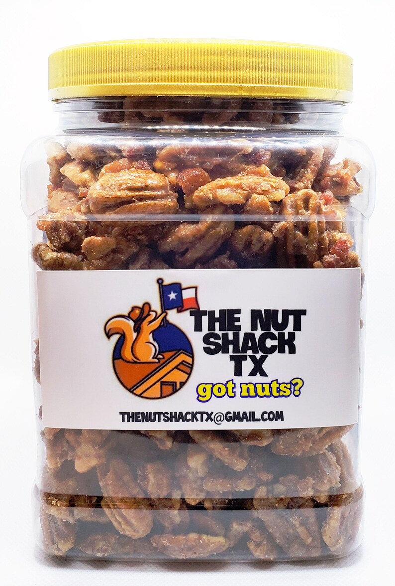 K&ouml;nnte beinhalten: Ein durchsichtiges Plastikglas gef&uuml;llt mit Pekannussh&auml;lften, bedeckt mit einem gelben Deckel. Das Glas hat ein wei&szlig;es Etikett mit dem Text "THE NUT SHACK TX got nuts?" und einem Logo eines Eichh&ouml;rnchens, das eine Flagge h&auml;lt.