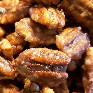 Puede incluir: Primer plano de nueces pecanas glaseadas, mostrando un recubrimiento brillante de color marr&oacute;n dorado. Las nueces est&aacute;n apiladas, resaltando su textura natural y su rico color. Un delicioso manjar.