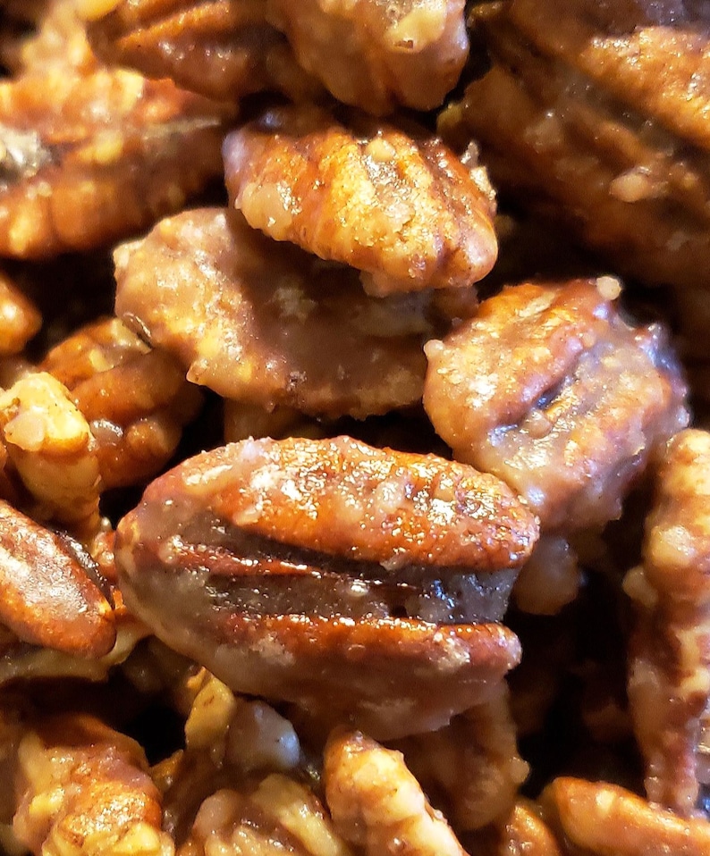 Puede incluir: Primer plano de nueces pecanas glaseadas, mostrando un recubrimiento brillante de color marr&oacute;n dorado. Las nueces est&aacute;n apiladas, resaltando su textura natural y su rico color. Un delicioso manjar.