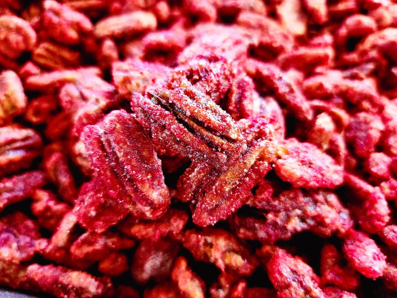 K&ouml;nnte beinhalten: Nahaufnahme von kandierten Pekann&uuml;ssen, die eine satte, r&ouml;tlich-braune Farbe mit einer Zuckerschicht aufweisen. Die N&uuml;sse sind eng aneinandergereiht und f&uuml;llen das Bild aus. Das Bild hebt die Textur und S&uuml;&szlig;e des Snacks hervor.