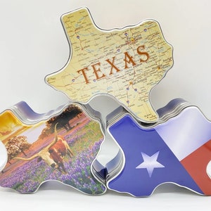 K&ouml;nnte beinhalten: Drei Texas-f&ouml;rmige Dosen. Eine Dose zeigt eine Karte von Texas mit dem Wort "TEXAS". Eine andere zeigt die Texas-Flagge. Die dritte Dose zeigt einen Longhorn in einem Feld mit Lupinen. Die Dosen sind aus Metall.