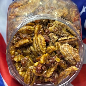Puede incluir: Un tarro de pl&aacute;stico transparente lleno de nueces pecanas confitadas. Las nueces pecanas son una mezcla de marr&oacute;n claro y oscuro, con un recubrimiento brillante. Algunas piezas tienen un tono rojizo. El tarro est&aacute; sobre un fondo de bandera tricolor.