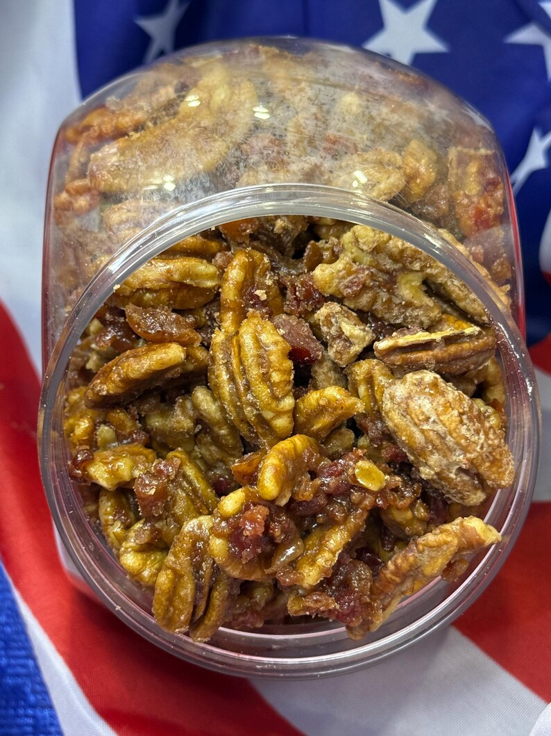 Puede incluir: Un tarro de pl&aacute;stico transparente lleno de nueces pecanas confitadas. Las nueces pecanas son una mezcla de marr&oacute;n claro y oscuro, con un recubrimiento brillante. Algunas piezas tienen un tono rojizo. El tarro est&aacute; sobre un fondo de bandera tricolor.