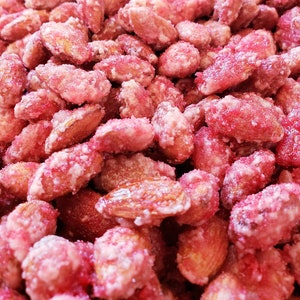 May include: Close-up of sugared almonds. The almonds are coated in a vibrant red sugar, with a dusting of white sugar crystals. The nuts are a light brown color, and the image is well-lit, highlighting the texture and color of the candy.