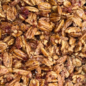 Puede incluir: Primer plano de nueces pecanas confitadas con un recubrimiento brillante y caramelizado. Las nueces son de un color marr&oacute;n intenso, con texturas y formas visibles. Peque&ntilde;os trozos de fruta roja seca est&aacute;n intercalados.