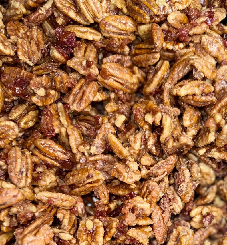 Puede incluir: Primer plano de nueces pecanas confitadas con un recubrimiento brillante y caramelizado. Las nueces son de un color marr&oacute;n intenso, con texturas y formas visibles. Peque&ntilde;os trozos de fruta roja seca est&aacute;n intercalados.